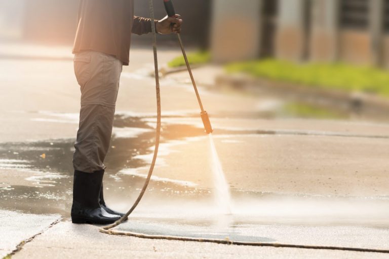 A worker power washing a concrete driveway. The high-pressure spray cleans grime, highlighting driveway cleaning effectiveness. Boots, hose, and a sunlit background are visible.