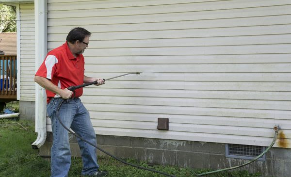 Man pressure washing vinyl siding of a house, cleaning away dirt and grime. He is wearing a red shirt and jeans, and the house has light-colored siding and a dark foundation.