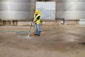 Worker in hard hat pressure washing a concrete surface in Columbia, MD. He's using a pressure washer to clean near large metal tanks, highlighting pressure washing Columbia services.