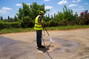 A worker in a yellow vest and hard hat uses a power washer on an asphalt road. He's performing power washing in Baltimore MD, removing stains from the pavement on a sunny day.