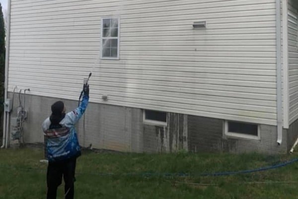 A man power washes the white siding of a residential house. He wears a blue jacket and black pants while spraying the wall, with a green lawn in the foreground.