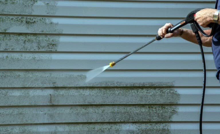 House washing in progress: a person pressure washing siding covered in green algae. The siding is half clean, showcasing the before and after of the house washing process.