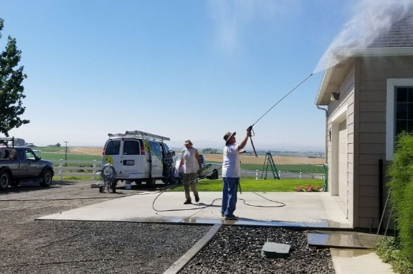 Two workers perform residential power washing on a light beige house under a clear blue sky. A white van and truck are parked nearby on a gravel driveway, with green fields in the background.